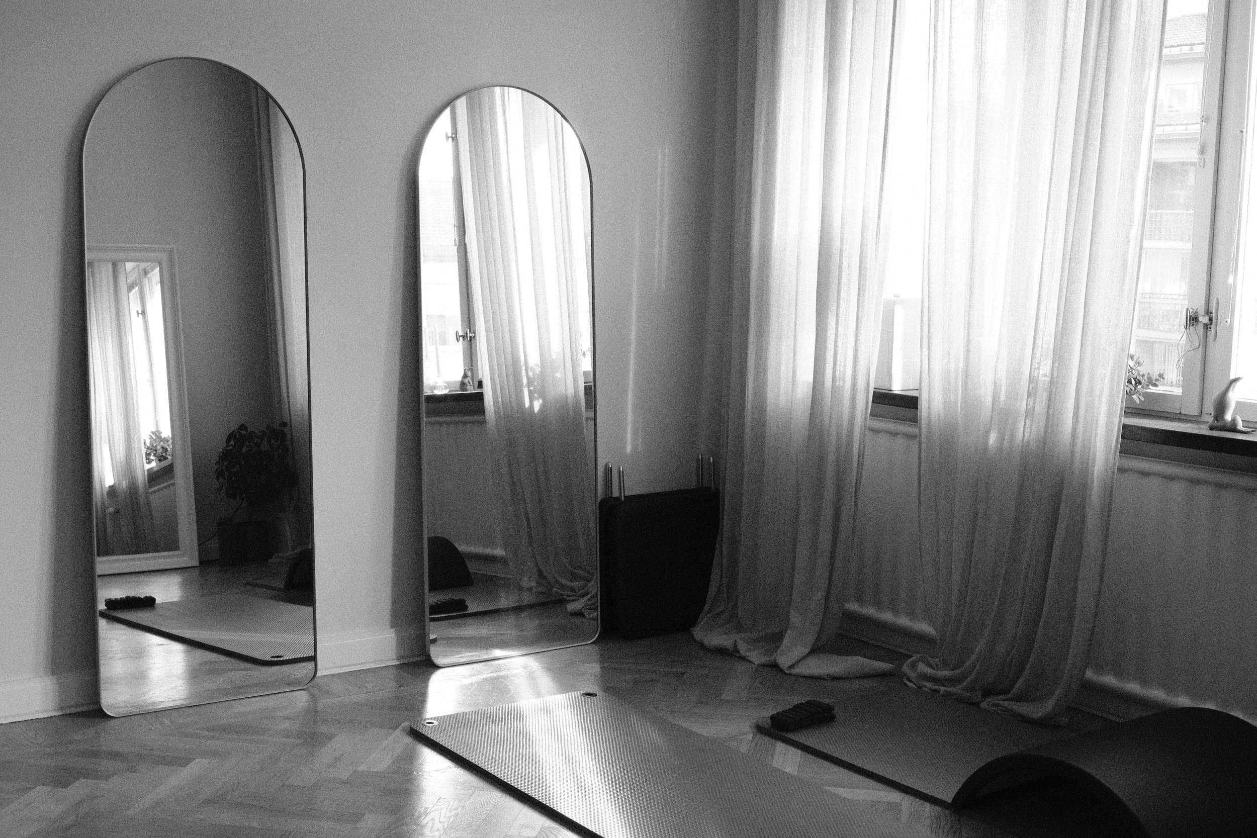 Black and white: the studio's two tall arched mirrors beside sheer curtains, mats and an arc barrel on the floor.