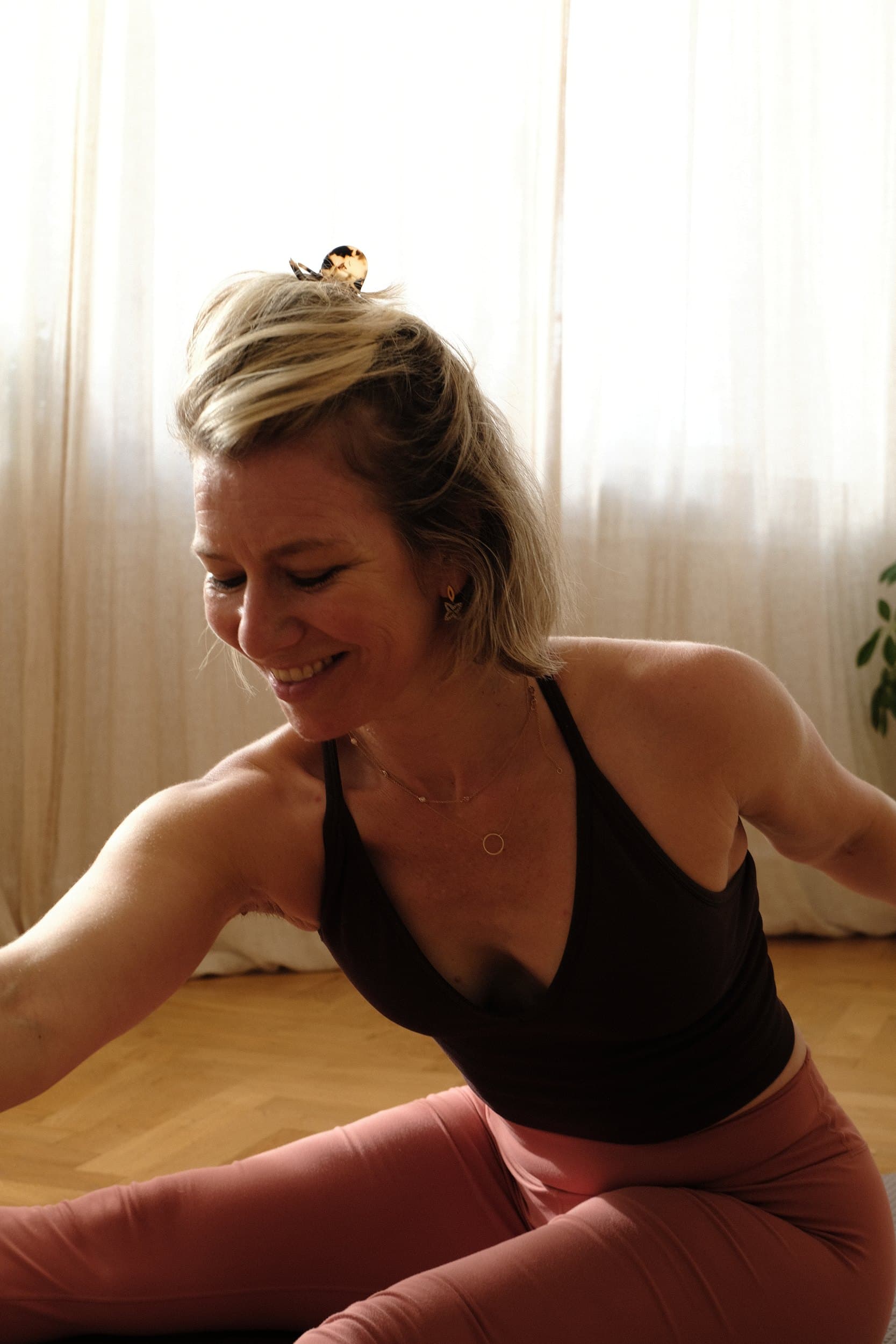 Liv, smiling, seated on her mat with her arms open in the soft window light of her home studio.